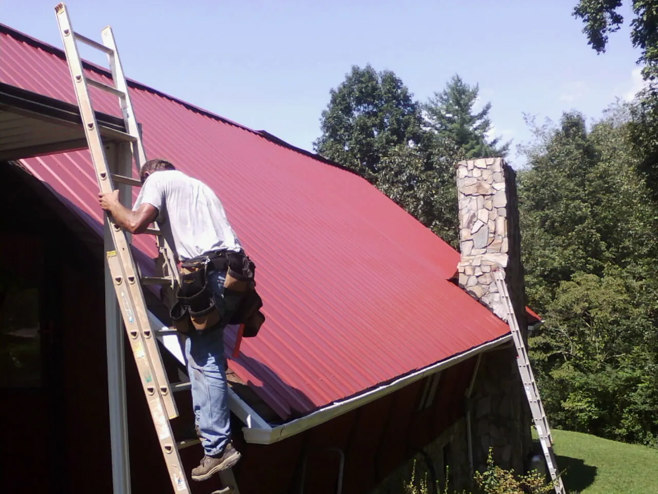 Roofer on a ladder beside a red metal roof and stone chimney — Soffit & Fascia Repair by Roofing Brothers in Alstead